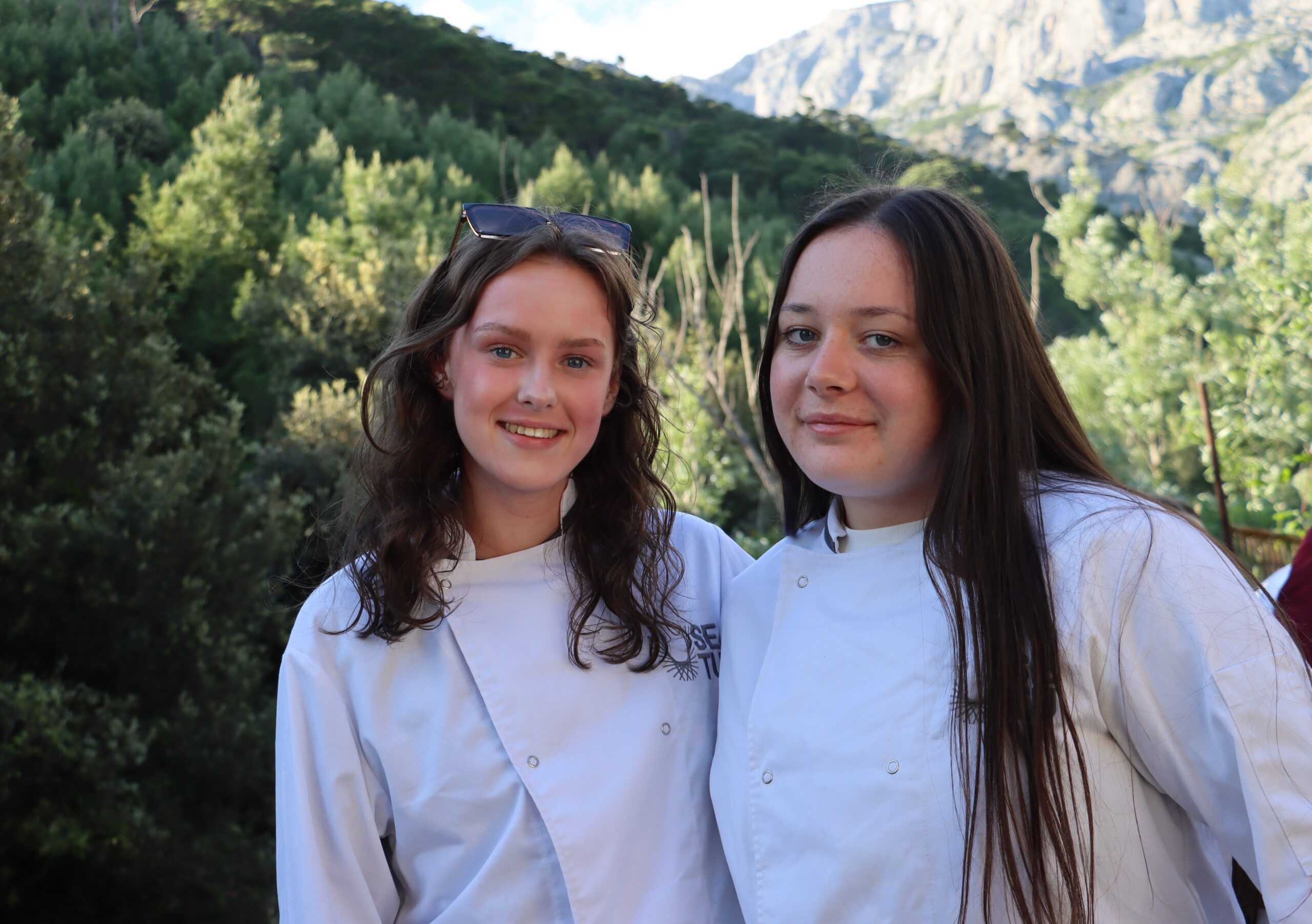 Two female students in their chef uniform. High green trees in the background 